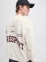 Buzo de manga larga color blanco roto (sea salt) con cuello redondo y calce oversize cropped. Presenta un estampado gráfico en la espalda con texto en color borgoña, incluyendo las palabras "RESPORT", "ATHLETIC ESSENTIALS", "CREATIVE DEPT." y el año "1990".