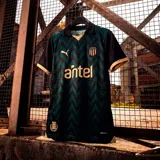 Camiseta de fútbol Puma del Club Atlético Peñarol, modelo Third Jersey 26. Diseño en tonos negro y verde con patrón en zigzag, detalles en dorado en cuello y mangas, logo de Puma, escudo del club y sponsor Antel en el frente.