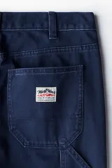 Pantalón tipo worker confeccionado en sarga de algodón de color azul marino, con un corte relaxed fit y piernas rectas. Presenta una cintura clásica con cierre de botón, bolsillos laterales al bies, bolsillos traseros, una trabilla funcional en un lateral y un bolsillo cargo dividido en el otro.