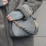 Cartera bandolera color gris de cuero sintético con diseño acolchado. Tiene correa regulable, cierre principal y dos compartimentos internos con bolsillos.