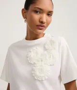 Blusa boxy de manga corta, confeccionada en algodón, con cuello redondo y detalles de flores de tejido aplicadas en el hombro y sisa.