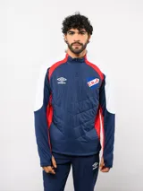 Buzo de entrenamiento Umbro del Club Nacional de Football, con cierre frontal parcial y cuello alto. Presenta un diseño con bloques de color azul, blanco y rojo, con panel frontal acolchado y mangas largas con aberturas para pulgares.