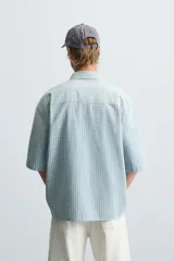 Camisa de corte relajado confeccionada en tejido de algodón, con estampado de cuadros pequeños en tonos celestes y blancos. Presenta cuello solapa, manga corta y bolsillo plastrón en el pecho con cierre frontal de botones.