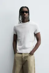 Camiseta de corte relajado, color gris claro, confeccionada en tejido con estructura waffle. Presenta cuello redondo y manga corta, con costuras decorativas en contraste color dorado que recorren los laterales y los hombros.