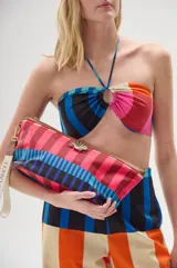 Necessaire de lona estampada con diseño de rayas multicolor. Presenta detalles en cuero ecológico, cierre superior metálico, aplique decorativo en forma de caracola dorada y correa lateral para llevar en la mano.
