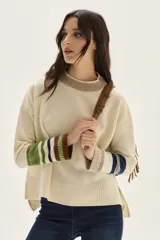 Sweater tejido de lambswool color beige, con cuello redondo y puños acanalados con rayas en tonos verdes, azules y marrones. Presenta flecos decorativos en los laterales de las mangas y aberturas en los costados.