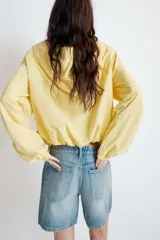 Chaqueta de nylon color amarillo pastel, con cuello subido y capucha. Presenta cierre frontal con cremallera, mangas largas con puños elásticos y detalle de lazadas ajustables en el frente. El bajo cuenta con cordón elástico y stoppers para un ajuste personalizado.