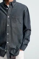 Camisa de corte relajado confeccionada en tejido de algodón denim, con cuello solapa, manga larga con puños abotonados y bolsillo plastrón en el pecho. Presenta cierre frontal de botones y bajo con aberturas laterales.