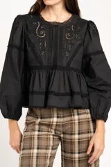 Blusa negra de algodón con diseño de broderie y calados en el pecho y hombros. Presenta corte oversize, mangas largas con puños y un detalle de volado en la cintura.