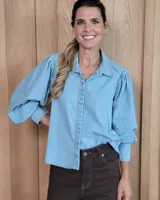 Camisa de jean en tono celeste claro, con cuello clásico, cierre frontal de botones y mangas largas abullonadas con puños ajustados.