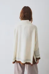 Buzo tejido de silueta amplia con cuello alto, mangas largas y puños en contraste. Presenta un diseño con franjas horizontales en el ruedo y detalles de tejido trenzado en las mangas.