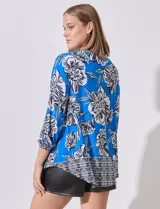 Blusa de crepe estampada con flores blancas y negras sobre fondo azul. Tiene escote entrelazado con abertura, mangas 3/4 con puños elastizados y corte holgado.