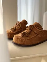 Zueco tipo mocasín destalonado confeccionado en gamuza color tabaco, con detalle de cordones y borlas en el empeine y costura reforzada en el contorno de la puntera.