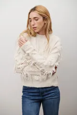 Buzo de punto color crema, con cuello redondo, manga larga y flecos decorativos en las mangas y el bajo.