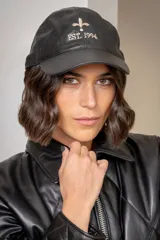 Gorra tipo baseball de cuero, color negro, con bordado frontal en hilo color crema que incluye una flor de lis y la inscripción 'EST. 1994'.