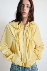 Chaqueta de nylon color amarillo pastel, con cuello subido y capucha. Presenta cierre frontal con cremallera, mangas largas con puños elásticos y detalle de lazadas ajustables en el frente. El bajo cuenta con cordón elástico y stoppers para un ajuste personalizado.