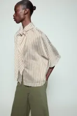 Camisa de corte holgado confeccionada en mezcla de lyocell, con estampado de rayas verticales en tonos neutros y azules. Presenta cuello redondo con lazada frontal, cierre con botones ocultos y mangas amplias por debajo del codo.