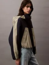 Campera corta con capucha, confeccionada en tejido ripstop de nailon en dos tonos: beige y negro. Tiene corte relaxed, cierre de cremallera y botón de presión, bolsillos con cremallera en la cintura, bajo con cordón elástico, forro de malla transpirable y logo CK bordado en el pecho.