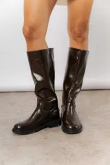 Bota de caña alta con acabado brillante, punta redondeada y suela plana. Presenta un detalle metálico decorativo en el lateral del tobillo.