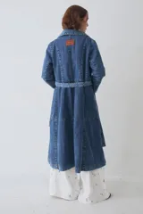 Trench largo de denim azul, con diseño de doble botonadura, solapas clásicas y mangas largas con puños ajustables. Presenta un corte recto y estructurado.