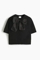 Saco de punto suave color negro, de manga corta y cuello redondo. Presenta cierre frontal con botones forrados y una moña decorativa de organza en la parte superior.