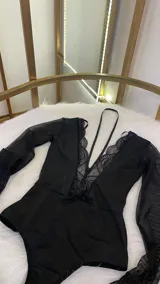 Body de lycra y encaje color negro, con mangas largas de tul transparente y detalle de encaje en los puños. Presenta un escote profundo con detalle de encaje y un collarín integrado que se conecta al centro del pecho.
