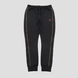 Pantalon de jogging Umbro color negro, con cintura elástica con cordón ajustable, puños acanalados en los tobillos y detalles de líneas verticales en color naranja a lo largo de las piernas. Presenta el logo de la marca bordado en color naranja en la parte superior.