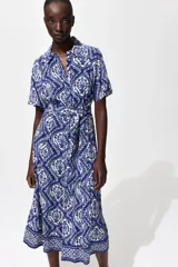 Vestido midi camisero de color azul con estampado ornamental en tonos blancos y azules. Cuenta con mangas cortas raglán, botonadura frontal completa y cinturón de la misma tela para atar en la cintura.
