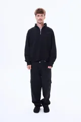 Buzo de felpa color negro con cuello alto y cierre frontal parcial. Presenta un diseño holgado con bolsillos laterales y logo bordado en el pecho.