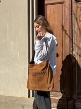 Cartera tipo tote bag de cuero color camel, con detalles de tachas metálicas en el bolsillo frontal y tiras largas para el hombro.