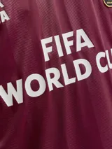Camiseta de estilo deportivo con cuello polo en color blanco y cuerpo en color bordó con diseño de rayas verticales sutiles. Presenta estampados frontales con la inscripción 'FIFA WORLD CUP' y 'CAN MEX USA', junto con un parche conmemorativo de la FIFA.