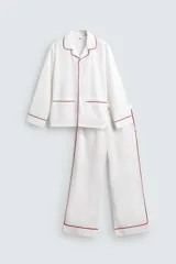 Pijama de dos piezas confeccionado en popelín de algodón blanco, con vivos de ribete en color bordó. Incluye una camisa de manga larga con cuello bowling, bolsillos de plastrón y cierre frontal de botones, y un pantalón de corte relajado con cintura elástica y bolsillos laterales.