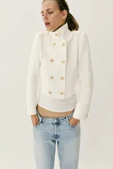 Chaqueta corta blanca de estilo militar, con cuello subido, manga larga y trabillas desflecadas en los hombros. Cuenta con cierre frontal cruzado decorado con botones metálicos dorados.
