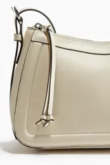 Cartera de hombro color crema, con diseño estructurado, cierre superior con tirador largo y correa ajustable con hebillas metálicas laterales.