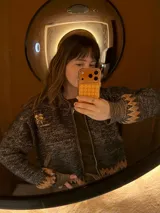 Saco tipo bomber de tejido jacquard en color negro con detalles en tono bronce. Presenta cierre frontal de aluminio, puños y ruedo con diseño geométrico en zigzag, y un bordado decorativo en el pecho.