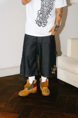 Bermuda de jean color azul oscuro (raw), corte baggy carpintero, con grandes bolsillos frontales y traseros. Presenta un bordado dorado con un monograma en la parte inferior lateral derecha.