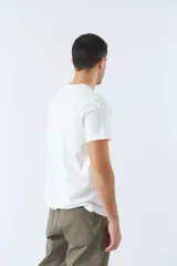 Remera blanca de algodón con cuello redondo y mangas cortas. Estampado frontal con diseño rectangular en tonos azules que incluye texto, coordenadas y la imagen de un avión.