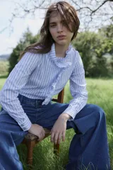 Camisa de algodón con estampado de rayas verticales en blanco y celeste. Presenta un diseño de manga larga con puños abotonados, cuello alto y un detalle de volado frontal que recorre la abertura de la prenda.