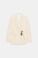 Blazer de corte estructurado en color beige, con cuello solapa y manga larga. Presenta un diseño cruzado con cierre mediante lazada del mismo tejido y un distintivo aplique metálico dorado en el frente.