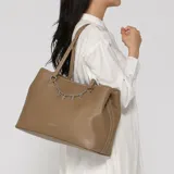 Cartera tipo tote Guess modelo Angelica, color marrón símil cuero, con doble asa superior y cadena colgante con dijes plateados que forman el nombre de la marca. Cuenta con logo impreso en la parte frontal y herrajes plateados.