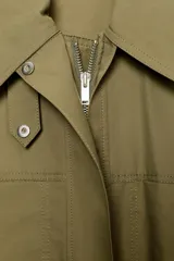 Vestido corto verde militar de cuello solapa y manga larga, con bolsillos de plastrón en el delantero y cierre frontal con cremallera oculta por sopata. Cuenta con un cinturón incorporado del mismo tejido con hebilla metálica y aberturas laterales en el bajo.