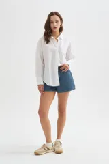 Camisa blanca de algodón, de corte holgado, con cuello camisero, cierre frontal de botones y un bolsillo aplicado en el pecho.