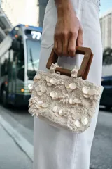 Bolso de mano formato mini confeccionado en tejido texturizado color crema. Presenta apliques de nácar y bordados florales en relieve, con doble asa rígida de madera y cadena bandolera extraíble.
