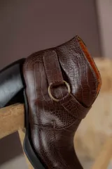 Bota corta de cuero vacuno con textura de reptil, color marrón oscuro. Cuenta con un taco cuadrado negro, suela de plataforma y una tira decorativa con hebilla metálica circular en el tobillo.