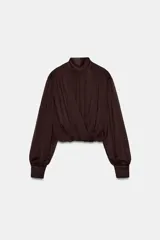 Blusa corta marrón de tejido fluido y semitransparente, con cuello subido, diseño cruzado drapeado en el frente y mangas largas abullonadas con puños ajustados.