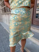 Conjunto de dos piezas compuesto por un top de un solo hombro con volados y una falda corta con ruedo asimétrico, confeccionado en tela satinada con estampado floral en tonos verde y dorado.