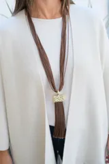 Collar largo compuesto por múltiples hilos finos de color marrón chocolate, con un dije central rectangular de metal dorado con textura irregular y un fleco largo del mismo tono que los hilos.