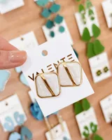 Aros de diseño geométrico cuadrado con acabado esmaltado en color blanco y detalles en metal dorado. Presentan una estructura metálica que divide la pieza en diagonal.