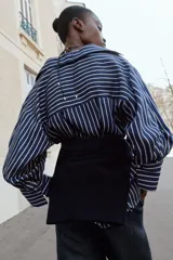 Camisa de rayas verticales azules y blancas, de cuello solapa con escote en V y cierre frontal con botones. Presenta mangas abullonadas por debajo del codo.