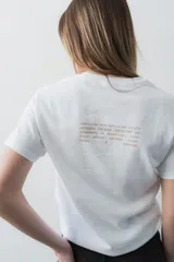 Remera de algodón blanca, con cuello redondo y mangas cortas. Presenta un estampado frontal con la palabra "KAPSUL" en rojo sobre un diseño floral en tono beige.